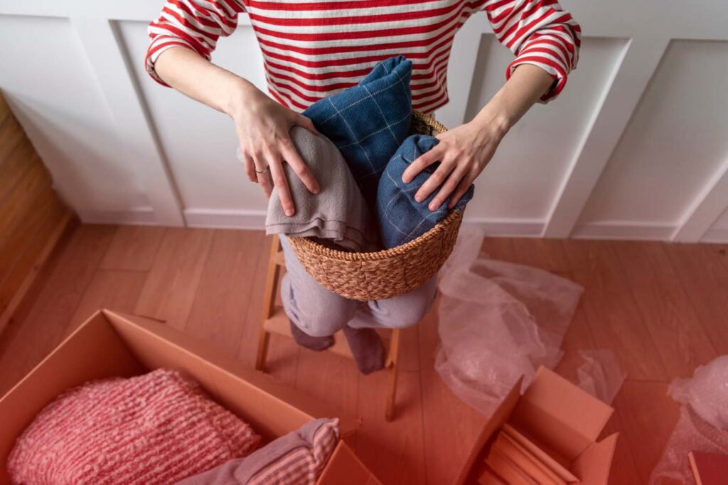 Person packing folded clothes into a basket surrounded by moving boxes during relocation.