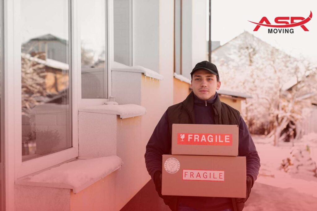 Mover carrying a fragile-labeled box outside a house during snowy winter conditions.