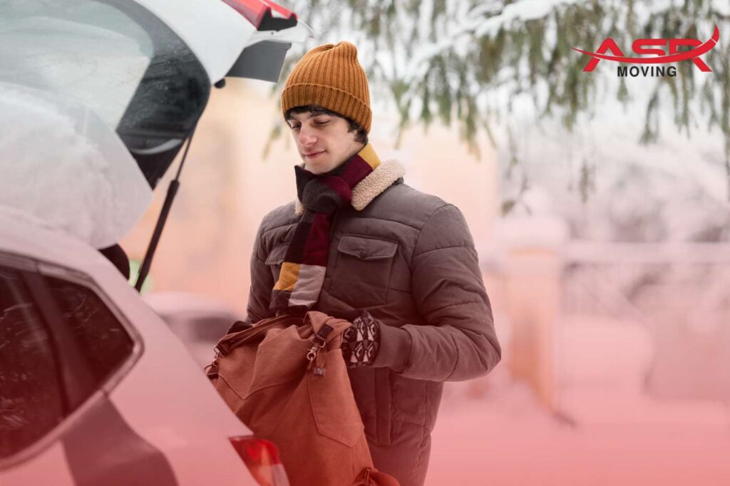 Person dressed in winter gear loading belongings into a car on a snowy moving day.